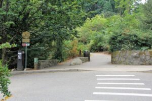 A crosswalk marked with white painted lines leading from a parking lot to the start of a wide paved trail framed by low stone walls to either side and abundant foliage from shrubby and tall trees all around it. A parking meter is to the left side of the crosswalk.