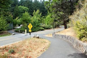 A wide paved trail with a low stone wall to the right and an area of dry grass to the left of that trail which is leading toward a crosswalk marked by a yellow sign. A combination of shrubby and tall trees both evergreen and broadleaf frame the edges of the image from the right and into the background.