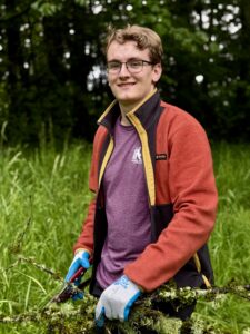A young man with short brown hair and glasses wearing a red, black, and yellow fleece, and gardening gloves holds pruning sheers and branches. He is looking directly at the camera and smiling.
