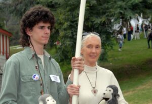 A young person with a dark curly mullet, a green button down shirt, and a slight smile, holding an ape-like stuffed animal holds a pole alongside famous biologist Jane Goodall who is also holding an ape-like stuffed animal and is wearing her silver hair pulled back, a white turtle neck, and an ornate necklace. They are standing in front of a grassy hill where people are gathering around a white tent.
