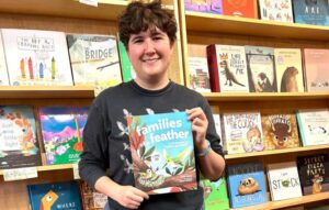 Fern Wrexler, weating a dark long-sleeved tshirt, and with dark short hair holds up their book Families of a Feather, looking and smiling directly to the camera. They are standing in front of a bookshelf lined with many childrens books.