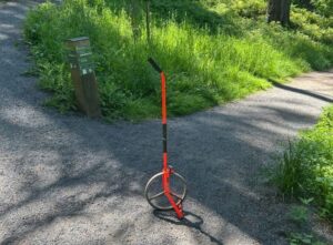 a wheel with and orange bar and black handle sits balanced at a trail intersection with an out of focus trail sign in the background surrounded by grass and tree trunks.