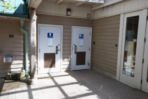 Two, closed bathroom doors are shown in the center of the frame with blue signs on them. The door on the left says "All User Restroom" with a white toilet icon, and the door on the right says "All User Restroom" with a toilet and wheelchair icon in white. There are water fountains shown to the left of the bathroom doors, and the front doors to the Visitor Center are shown on the right.