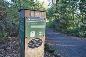 Trailhead of the Visitor Center Loop with a Trail Marker post in the foreground, showing the paved path and greenery along the trailsides.