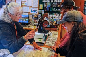 A woman stands behind a counter and provides instructions to 2 visitors on the other side of the counter by pointing to the trail map.