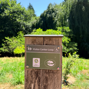 A wooden trail marker post at the corner of a trail above a sloping grassy hillside with "Visitor Center Loop" labeled at the top outlined in gray with the Hoyt Arboretum Friends and Portland Parks and Recreation logos underneath. A mix of tall broadleaf and conifer trees are in the background against a blue sky.