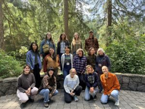 a group of 15 people of varying ethnicities and ages in 3 vertical layers smiling at the camera with a backdrop of tall evergreen trees.