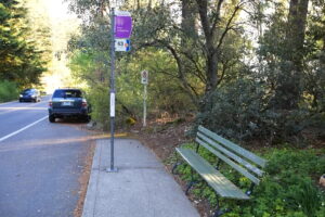 An image of the free shuttle stop in front of the Visitor Center. The stop is marked with a post and is next to a wooden bench.