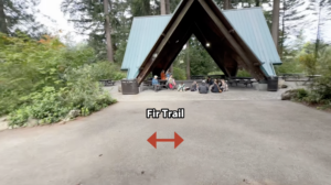 An image facing a green-roofed picnic pavilion surrounded by shrubbery and the wide paved entrance to the pavilion extending to a dirt trail on either side. The trail is labelled by a double headed red arrow pointing in either direction labelled with the words "Fir Trail". There are people gathered and sitting on the ground with young children at the Pavilion.