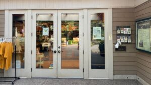 Glass front doors to a building with a t-shirt rack outside to the left, a brochure rack with maps to the right, and a bulletin display on the wall to the right of the doors. There is a sign in the front door that says "open".
