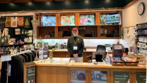 A smiling man wearing a red hat and a green long-sleeve t-shirt with a grey beard and glasses stands behind a desk with his hands clasped in front of him. He has a name tag on that reads "John". There are shirts, bags, gifts, and decorative tree images in the shop around him.