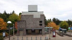 An image of a big grey building with an elevator, directional signs for the MAX Red-Blue Line and a public bus pulling up on the right.